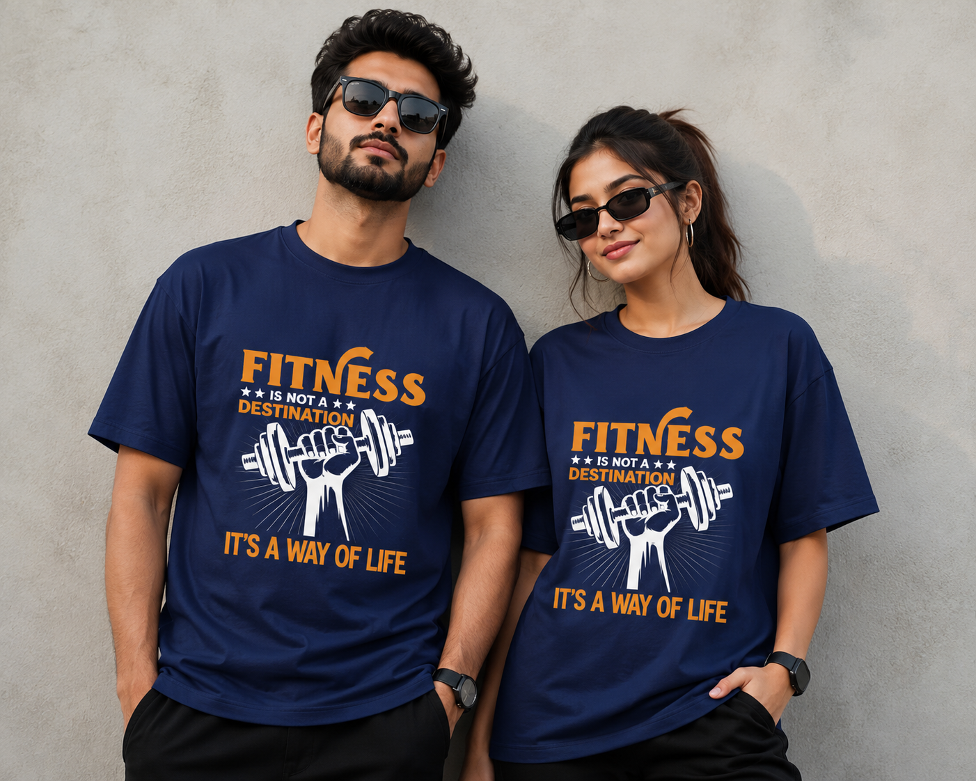 A man and a woman wearing matching navy blue t-shirts with a graphic print that reads 'FITNESS IS MY DESTINATION' and 'IT'S A WAY OF LIFE' in orange and white, standing against a textured gray wall.